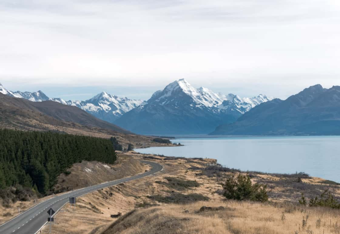 A winding road, dry grassland and lake with snow-capped mountains